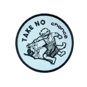 Op de afbeelding: Een ronde patch met een lichtblauwe achtergrond en zwarte rand. De patch heeft een zwart-wit illustratie van twee worstelende figuren. De woorden "TAKE NO chance" zijn in het zwart rond de rand van de patch gedrukt.