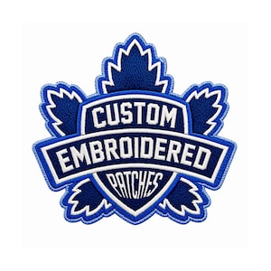 May include: A blue and white embroidered patch in the shape of a maple leaf. The patch features the text "CUSTOM EMBROIDERED PATCHES" in white lettering on a blue background. The design has a detailed, stitched appearance.