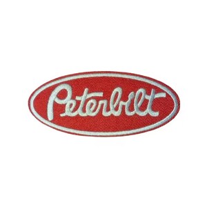 May include: A red, oval-shaped embroidered patch with the word "Peterbilt" in white script. The patch has a white border and is set against a white background. The patch is likely for a hat, jacket, or other apparel.