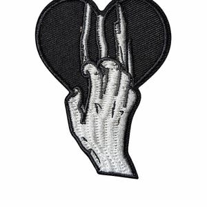 Può includere: Toppa ricamata raffigurante un cuore nero con una mano bianca che fa il segno della pace. La mano è dettagliata con dita visibili e un aspetto strutturato, su uno sfondo nero.