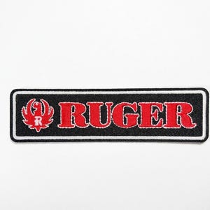 Pode incluir: Patch bordado retangular preto com a palavra "RUGER" em vermelho com contorno branco. Um logotipo vermelho est&aacute; presente &agrave; esquerda. Ideal para personalizar roupas e acess&oacute;rios.