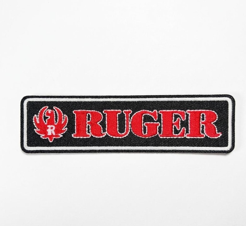 Pode incluir: Patch bordado retangular preto com a palavra "RUGER" em vermelho com contorno branco. Um logotipo vermelho est&aacute; presente &agrave; esquerda. Ideal para personalizar roupas e acess&oacute;rios.