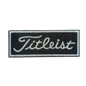 May include: A rectangular black patch with the word "Titleist" embroidered in white cursive script. The patch has a white border and is likely for apparel or accessories.
