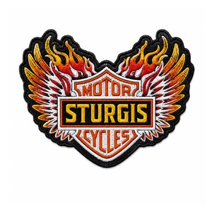 May include: An embroidered patch featuring a shield design with the text MOTOR STURGIS CYCLES in yellow and orange. The emblem is flanked by white and orange wings and topped with stylized orange and red flames, all outlined in black thread.