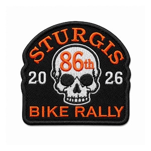 May include: A black embroidered patch featuring a white skull graphic in the center. The text reads STURGIS in orange, 86th in orange, 20 and 26 in white on the sides, and BIKE RALLY in orange. The patch has a black border and a curved top edge.