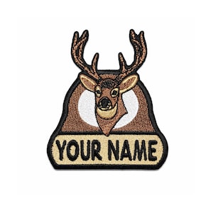 Könnte beinhalten: Gestickter Aufnäher mit einem Hirschkopf mit Geweih, vor einem braunen und weißen Hintergrund. Der Aufnäher enthält den Text "YOUR NAME" in Gold auf einem schwarzen Banner. Ideal für Jacken, Taschen oder andere Stoffartikel.