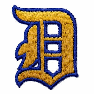 May include: Embroidered patch featuring a stylized letter "D" in gold with a blue outline and interior details. The patch has a classic, collegiate design, suitable for jackets, bags, or other fabric items. The letter has a bold, blocky font.