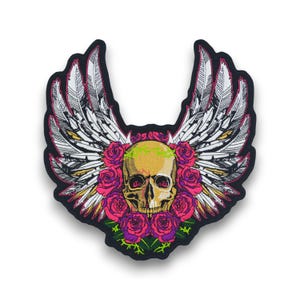Puede incluir: Parche negro con un cráneo dorado, rodeado de rosas rosas y alas blancas con plumas. El cráneo tiene ojos rojos y una corona verde. El parche tiene un borde negro.