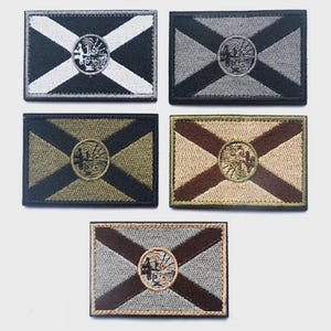 Può includere: Cinque toppe rettangolari ricamate con un disegno a bandiera. Ogni toppa presenta un emblema circolare centrale e un motivo a croce diagonale. Le combinazioni di colori includono bianco e nero, grigio, verde oliva, beige e marrone.