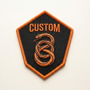 May include: A black and orange embroidered patch with the word "CUSTOM" at the top. The patch features an orange snake coiled in a figure-eight shape. The patch has an orange border.