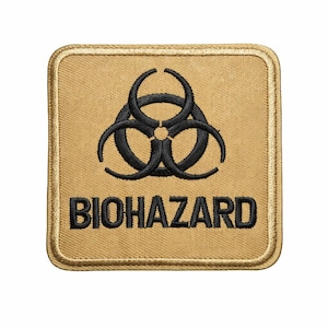 May include: A square, tan patch with a black biohazard symbol and the word "BIOHAZARD" embroidered in black. The patch has a gold border and is likely for clothing or gear.