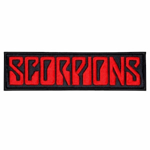 May include: A rectangular patch with the word "SCORPIONS" embroidered in red, bold, stylized letters. The text is set against a black background and framed by a black border. The patch is likely for clothing or accessories.