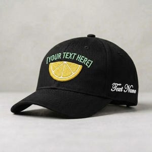 Puede incluir: Gorra de béisbol negra con una rodaja de limón bordada en la parte delantera. El texto "[YOUR TEXT HERE]" está encima del limón, y "Text Name" está bordado en el lateral. La gorra tiene una visera curva.