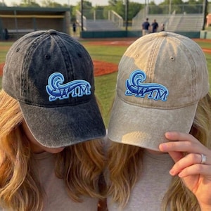 Könnte beinhalten: Zwei Baseballkappen, eine dunkelgraue und eine beige, jeweils mit einem blau-weißen Wellenmuster und dem Wort "SWIM" auf der Vorderseite gestickt. Die Kappen werden von Personen mit blondem Haar getragen, vor dem Hintergrund eines Baseballfeldes.