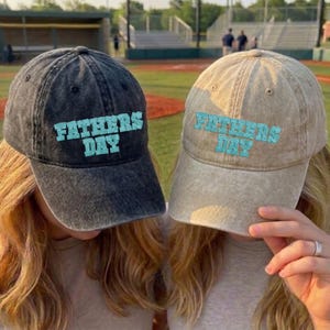 May include: Two baseball caps, one dark gray and one tan, with the text "FATHERS DAY" in turquoise. The caps are worn by two people, with the background of a baseball field.