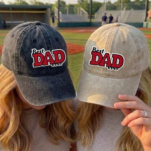 May include: Two baseball caps, one charcoal gray and one tan, each with the text "Best Dad Ever" in red and white. The caps are worn by two people, with the background of a baseball field.