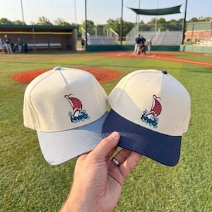 Könnte beinhalten: Zwei Baseballkappen mit cremefarbenem Oberteil und hellblauem bzw. marineblauem Schirm. Jede Kappe zeigt ein rot-weißes Segelboot-Logo über blauen Wellen. Die Kappen werden vor einem Baseballfeld gehalten.
