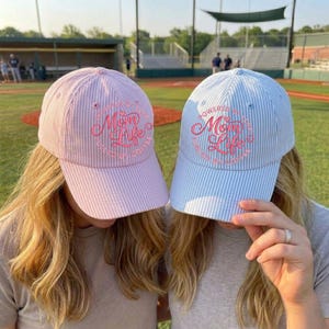 Puede incluir: Dos gorras de béisbol, una rosa y otra azul claro, con rayas verticales. La gorra rosa tiene el texto "Mom Life" en una fuente cursiva, y la gorra azul dice "Powered by Mom Life, Fueled by Coffee."