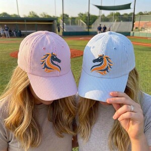May include: Two baseball caps, one pink and one light blue, with a seersucker stripe pattern. Each cap features an embroidered horse head logo in black, orange, and yellow. The caps are worn by two people in a baseball field setting.