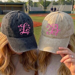 Op de afbeelding: Twee baseballpetten, één donkergrijs en één beige, worden getoond. Elke pet heeft een roze geborduurde initiaal met bloemmotieven. De petten worden door personen gedragen, met een honkbalveld op de achtergrond.
