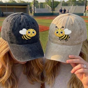 Peut inclure: Deux casquettes de baseball, une grise foncée et une beige, chacune ornée d'un écusson d'abeille de dessin animé. L'abeille a des rayures jaunes et noires, des ailes blanches et un visage souriant. Les casquettes sont portées par deux personnes.