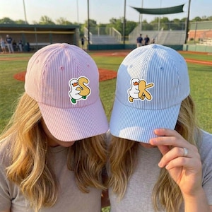 May include: Two baseball caps with a seersucker stripe pattern. One is pink with a duck and the letter "S" patch, the other is light blue with a duck and a baseball bat patch. Both are worn by people.
