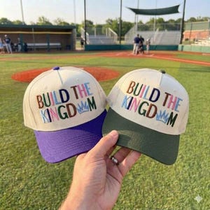 Può includere: Due cappellini da baseball con la scritta ricamata "BUILD THE KINGDOM". Uno ha la visiera viola, l'altro verde. Entrambi hanno il retro in rete color crema e il davanti bianco.