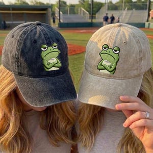 Puede incluir: Dos gorras de béisbol, una gris oscura y otra beige, cada una con un diseño de rana bordada. La rana tiene una expresión gruñona y sostiene una taza. Las gorras son usadas por dos personas, con un campo de béisbol de fondo.