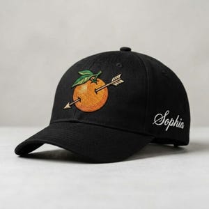 Könnte beinhalten: Schwarze Baseballkappe mit einer gestickten Orange, die von einem Pfeil durchbohrt wird, mit grünen Blättern. Der Name "Sophia" ist in weißer Schrift auf der Seite gestickt. Ein einzigartiges Accessoire.