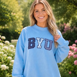 May include: A light blue long-sleeve sweatshirt featuring the letters BYU in a collegiate font across the chest. The letter B has a dark blue polka dot pattern, the Y has a light blue floral print, and the U has a dark blue gingham check pattern.