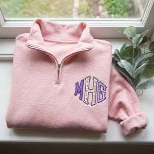 May include: A light pink quarter-zip pullover sweatshirt featuring a monogram on the left chest. The monogram consists of the letters MHG in a purple and white polka dot pattern and a yellow floral print with a dark purple outline.