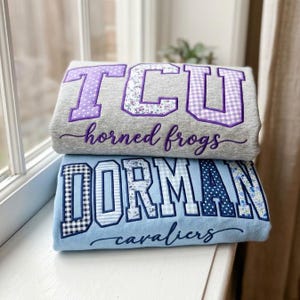 May include: Two folded sweatshirts stacked vertically. The top gray sweatshirt features purple appliqued letters reading TCU and purple cursive text reading horned frogs. The bottom light blue sweatshirt features navy blue letters reading DORMAN and navy cursive text reading cavaliers.