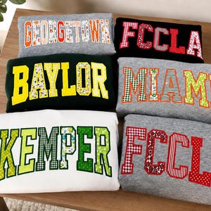 May include: Six folded sweatshirts in gray, black, white, and dark green. Each features large block-letter applique text including GEORGETOWN, FCCLA, BAYLOR, MIAMI, and KEMPER. The letters are filled with various patterns like gingham, floral, and polka dots.