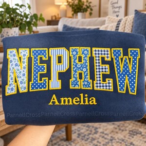 May include: A navy blue sweatshirt featuring the word NEPHEW in large, yellow-outlined block letters. Each letter is filled with a different blue and white pattern, including floral, polka dot, and striped designs. The name Amelia is embroidered in yellow below.