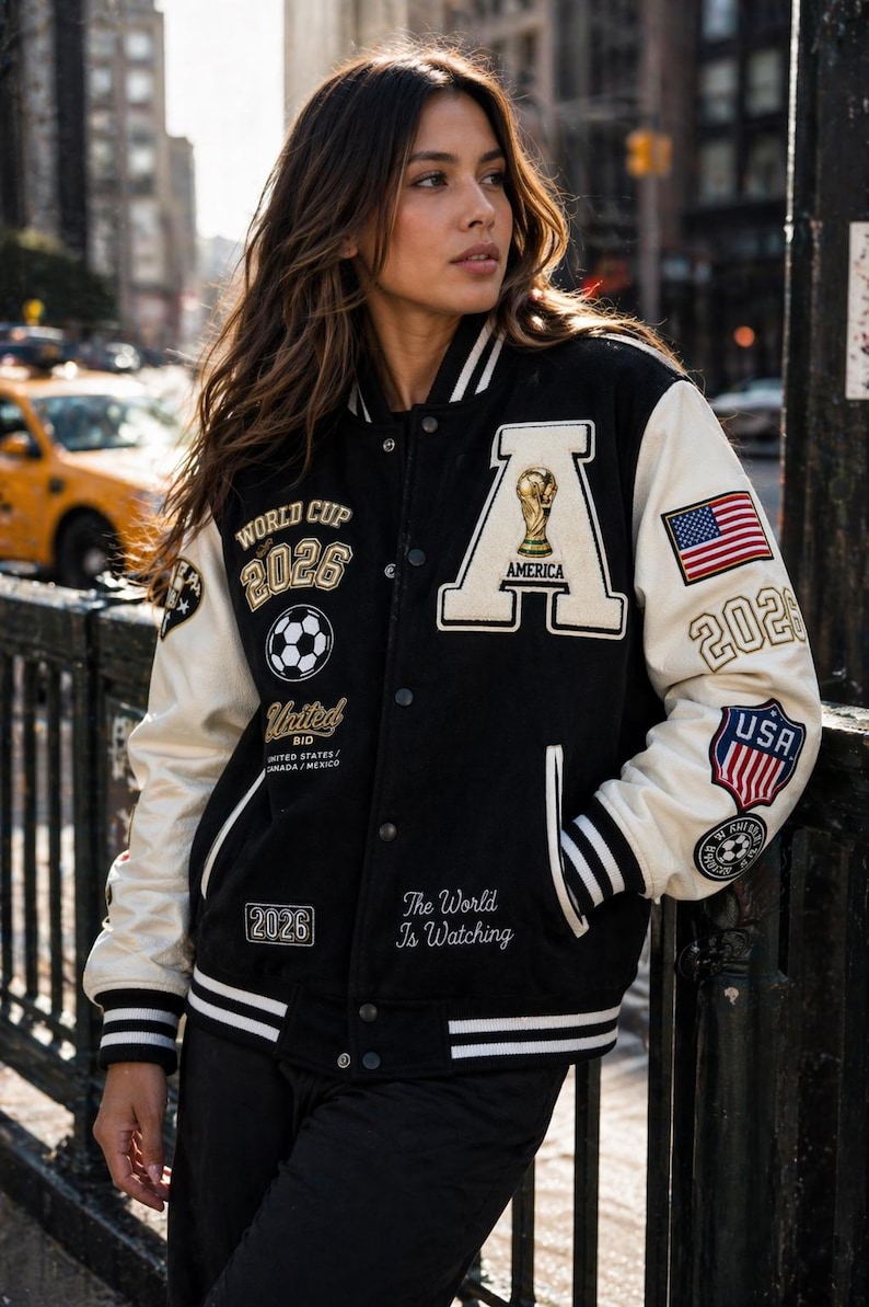 May include: Black and cream-colored letterman jacket with "WORLD CUP 2026" and "AMERICA" patches. Features a soccer ball and the text "The World Is Watching". Includes an American flag patch and the text "USA 2026".
