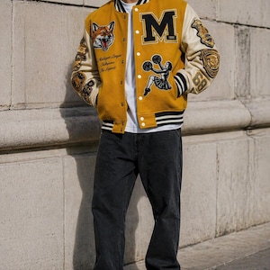 May include: Mustard yellow varsity jacket with black and cream accents. Features a large letter "M" and various patches, including a fox and cheerleader graphic. The jacket is worn with black jeans and white sneakers.