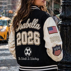 May include: Black and cream varsity jacket with the name "Isabella" and the year "2026" on the back. The jacket features a soccer ball graphic and the text "The World Is Watching." An American flag and "USA" emblem are on the sleeve.
