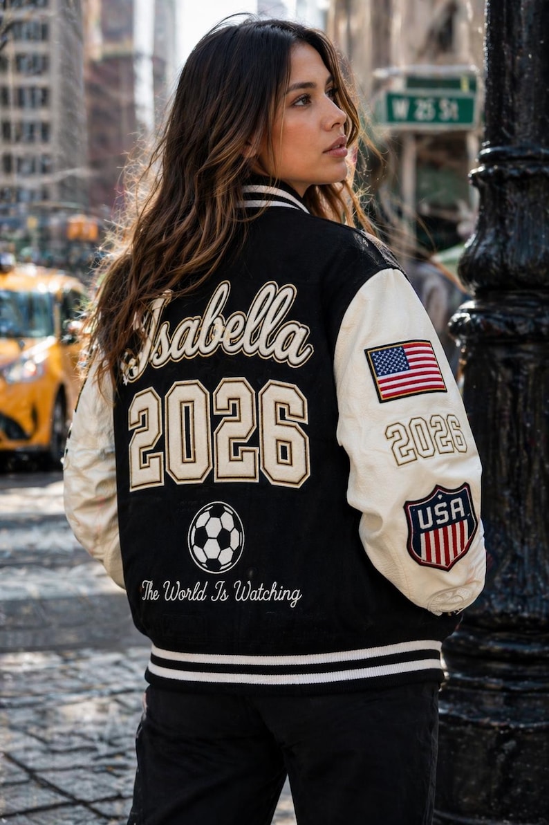 May include: Black and cream varsity jacket with the name "Isabella" and the year "2026" on the back. The jacket features a soccer ball graphic and the text "The World Is Watching." An American flag and "USA" emblem are on the sleeve.