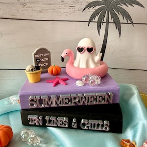 May include: A decorative stack of two books, one purple and one black, with the text "SUMMERWEEN" and "TAN LINES & CHILLS" on the spines. A white ghost in pink heart sunglasses sits in a pink flamingo floatie, with a palm tree, tombstone, and pumpkin.