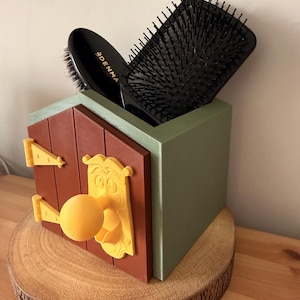 May include: A whimsical, 3D-printed brush holder in the shape of a door, with a brown door, yellow hinges and doorknob, and a green box. Two black hairbrushes are inside. The holder sits on a wooden slice.