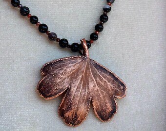 May include: A necklace featuring a copper-coloured leaf pendant with a textured, dark-toned finish. The pendant is suspended from a strand of small, round, black beads interspersed with tiny, dark red metallic accents along the necklace chain.