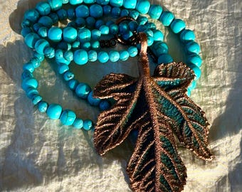 May include: A necklace featuring a large, textured copper-coloured leaf pendant with dark green accents, suspended from a strand of round, bright turquoise beads with dark veining. The piece includes a dark metal circular clasp connector.