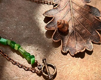 May include: A necklace featuring a large copper-coloured oak leaf pendant with a small snail shell attached to the surface. The chain is composed of green stone beads and copper-toned metal links, secured with a circular toggle clasp closure.