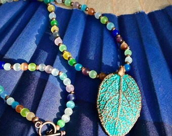 May include: A necklace featuring a series of multicoloured, translucent, round beads in shades of blue, green, yellow, and pink, accented with small copper-toned spacers. A large, textured, turquoise-coloured leaf pendant hangs from the centre.
