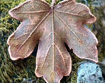 May include: A copper-toned metal leaf ornament featuring a textured, granular surface with prominent veins. The leaf has a distinct five-lobed shape with jagged edges, showcasing a metallic finish that transitions between reddish-brown and bronze hues.