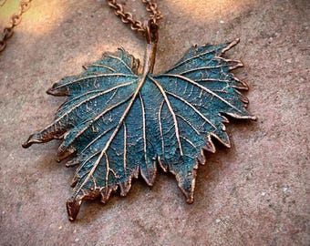May include: A necklace featuring a pendant shaped like a textured leaf with a deep teal and green patina finish. The leaf has prominent veins and jagged edges, suspended from a matching copper-toned metal chain with small circular links.