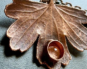 May include: A copper-toned metal pendant shaped like a textured maple leaf, featuring a small circular bezel setting containing a clear, round gemstone. The pendant is attached to a matching copper-coloured metal chain with visible links.