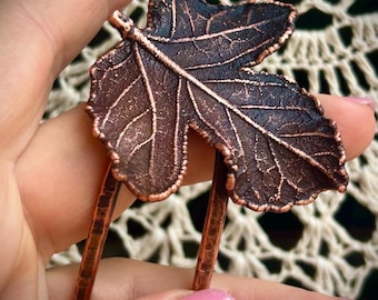 May include: A copper hair fork featuring a detailed, textured fig leaf design. The metal has a dark, oxidised finish with bright copper highlights along the edges and veins of the leaf. The two prongs are hammered and have a rustic, metallic appearance.