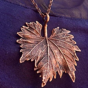 Op de afbeelding: Een koperkleurige metalen hanger in de vorm van een gedetailleerd druivenblad, met prominente nerven en een textuurrijk oppervlak. De bladvormige hanger hangt aan een bijpassende koperkleurige metalen ketting met een natuurgetrouw ontwerp.