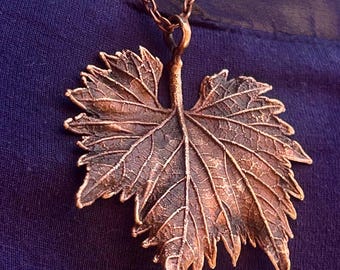 May include: A copper-toned metal pendant shaped like a detailed grape leaf, featuring prominent veins and a textured surface. The leaf pendant is suspended from a matching copper-coloured metal chain, showcasing a naturalistic design with jagged edges.